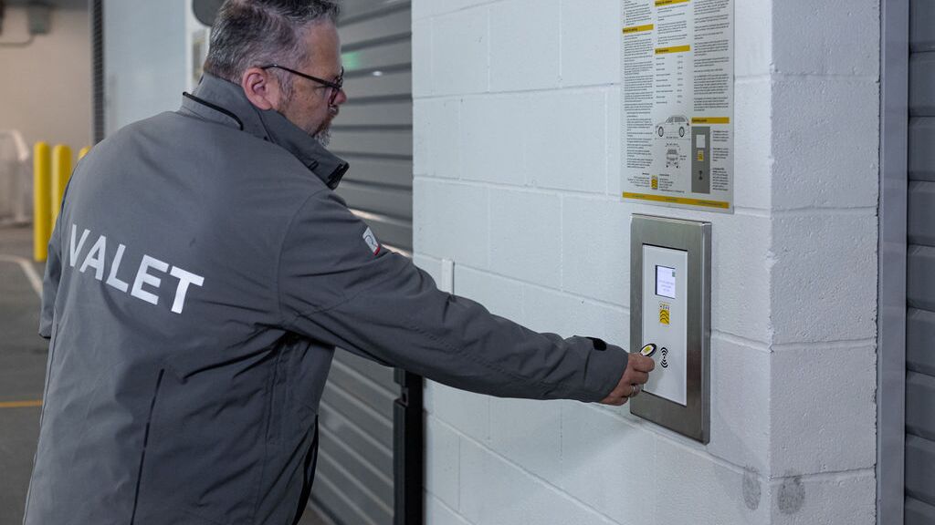 The image shows a person at the control panel of an automated parking system, confirming the storage process and thereby starting the automatic transport of the vehicle to an available parking space.