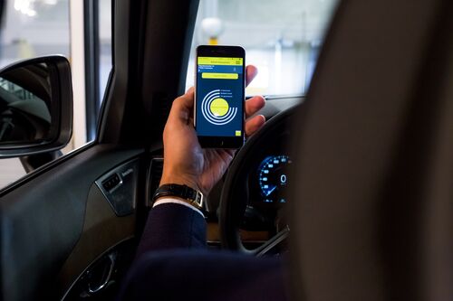 Une femme est assise dans sa voiture et tient devant elle un téléphone portable sur lequel une application est ouverte